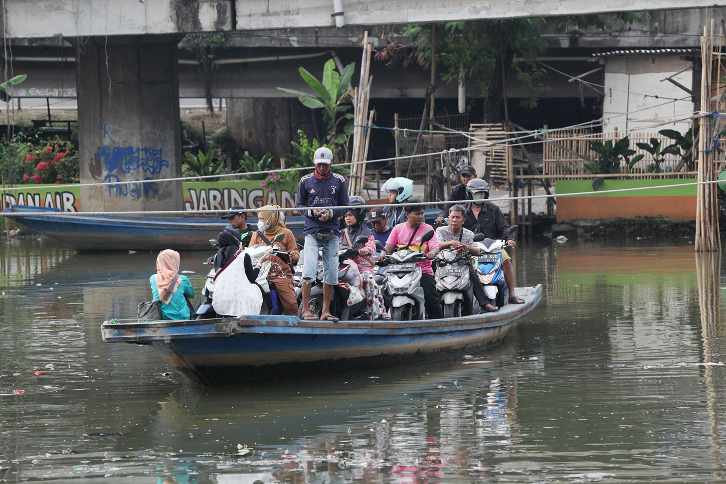 Sejumlah pengendara sepeda motor menggunakan perahu eretan menyeberangi Sungai Kali Angke di Kecamatan Penjaringan, Jakarta, Kamis (30/5/2024). (BeritaNasional.Com/Oke Atmaja)