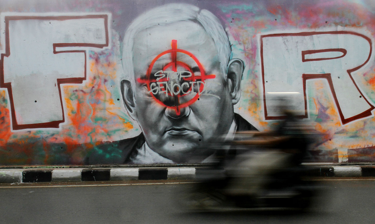 Pengendara sepeda motor melintasi mural dengan gambar wajah PM Israel, Benjamin Netanyahu dengan tulisan Stop Genocide di Kawasan Sudirman, Jakarta, Jumat(21/6/2024).(BeritaNasional.Com/Oke Atmaja)