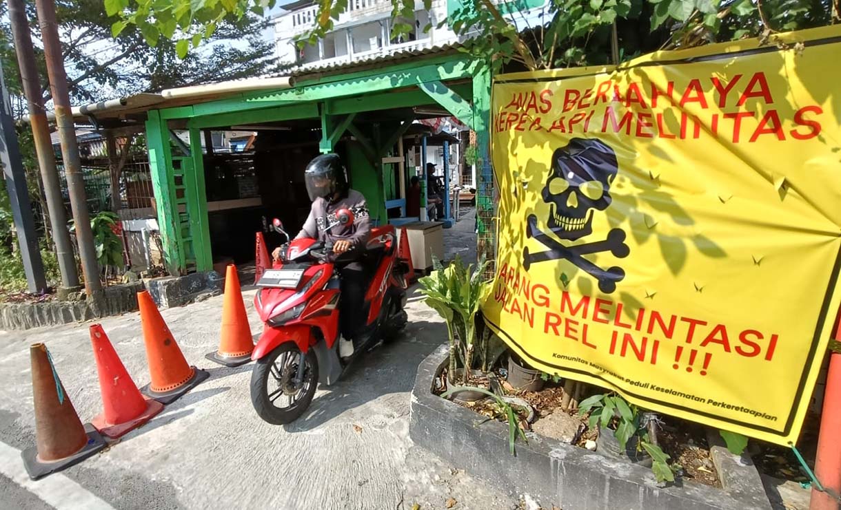 Pengendara melintas di samping spanduk peringatan dan larangan melintas di perlintasan liar jalur kereta api di kawasan Tebet, Jakarta, Selasa (19/5/2026).  (Beritanasional.com/Oke Atmaja)
