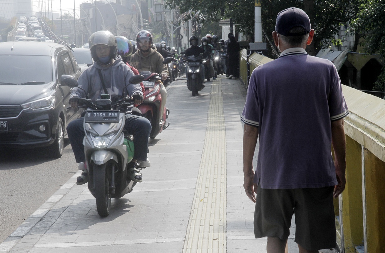 Warga berjalan di dekat pengendara motor yang melintasi trotoar di Jalan Matraman Raya, Jakarta, Selasa (26/4/2024).(Indonesiaglobe/Oke Atmaja)