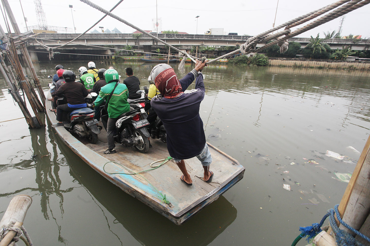 Sejumlah pengendara sepeda motor menggunakan perahu eretan menyeberangi Sungai Kali Angke di Kecamatan Penjaringan, Jakarta, Kamis (30/5/2024). (BeritaNasional.Com/Oke Atmaja)