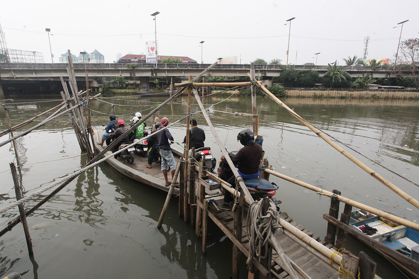 Sejumlah pengendara sepeda motor menggunakan perahu eretan menyeberangi Sungai Kali Angke di Kecamatan Penjaringan, Jakarta, Kamis (30/5/2024). (BeritaNasional.Com/Oke Atmaja)