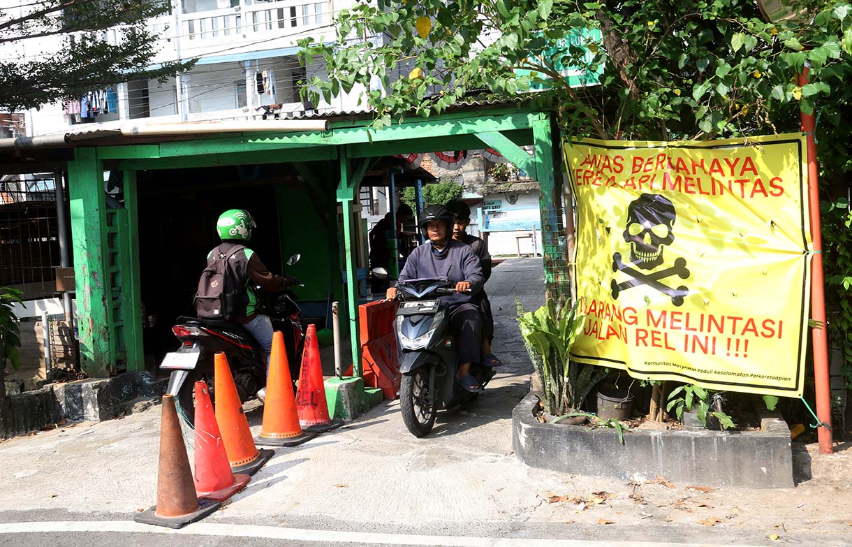 Pengendara melintas di samping spanduk peringatan dan larangan melintas di perlintasan liar jalur kereta api di kawasan Tebet, Jakarta, Selasa (19/5/2026).  (Beritanasional.com/Oke Atmaja)
