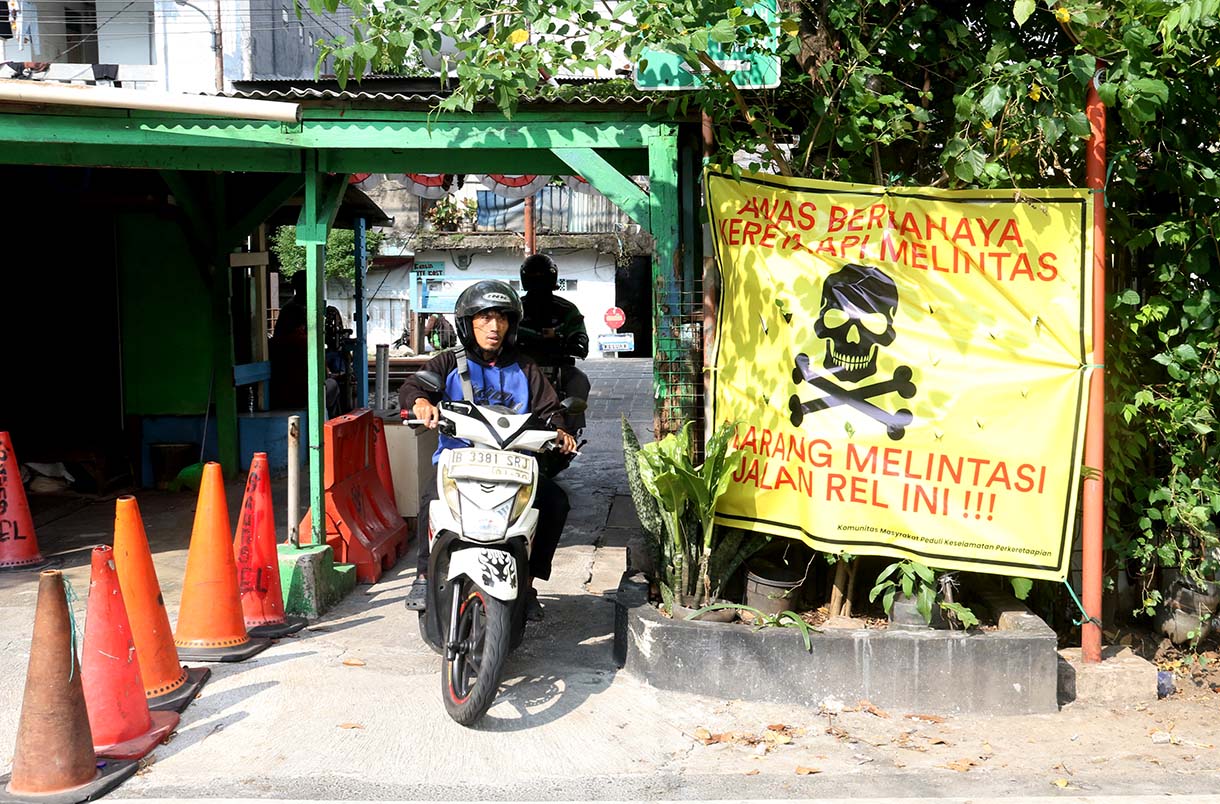 Pengendara melintas di samping spanduk peringatan dan larangan melintas di perlintasan liar jalur kereta api di kawasan Tebet, Jakarta, Selasa (19/5/2026).  (Beritanasional.com/Oke Atmaja)
