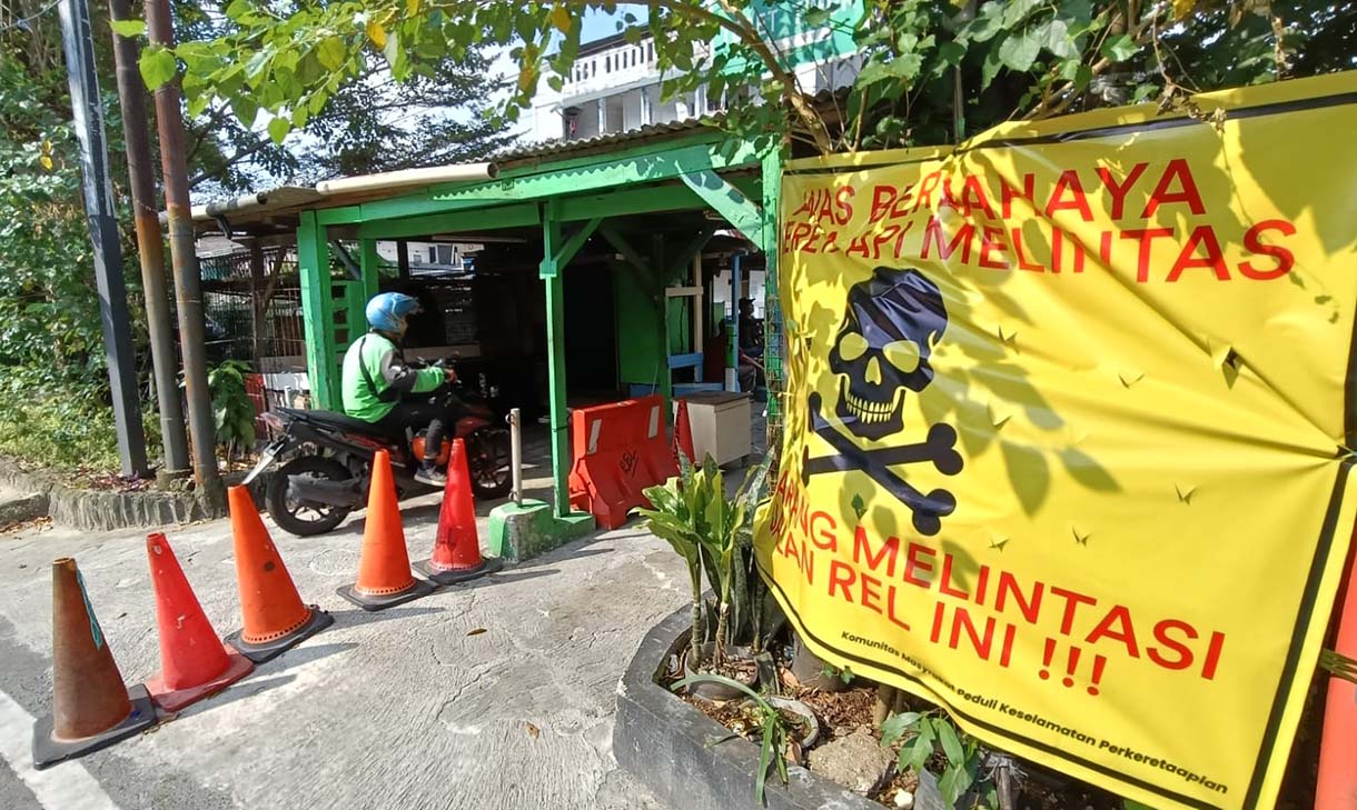 Pengendara melintas di samping spanduk peringatan dan larangan melintas di perlintasan liar jalur kereta api di kawasan Tebet, Jakarta, Selasa (19/5/2026).  (Beritanasional.com/Oke Atmaja)